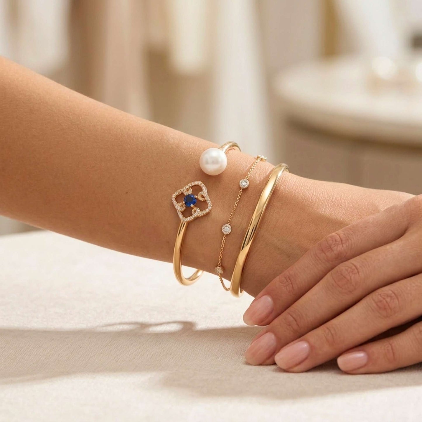 Close-up of a wrist wearing multiple gold bracelets with pearls and gemstones on a neutral background.