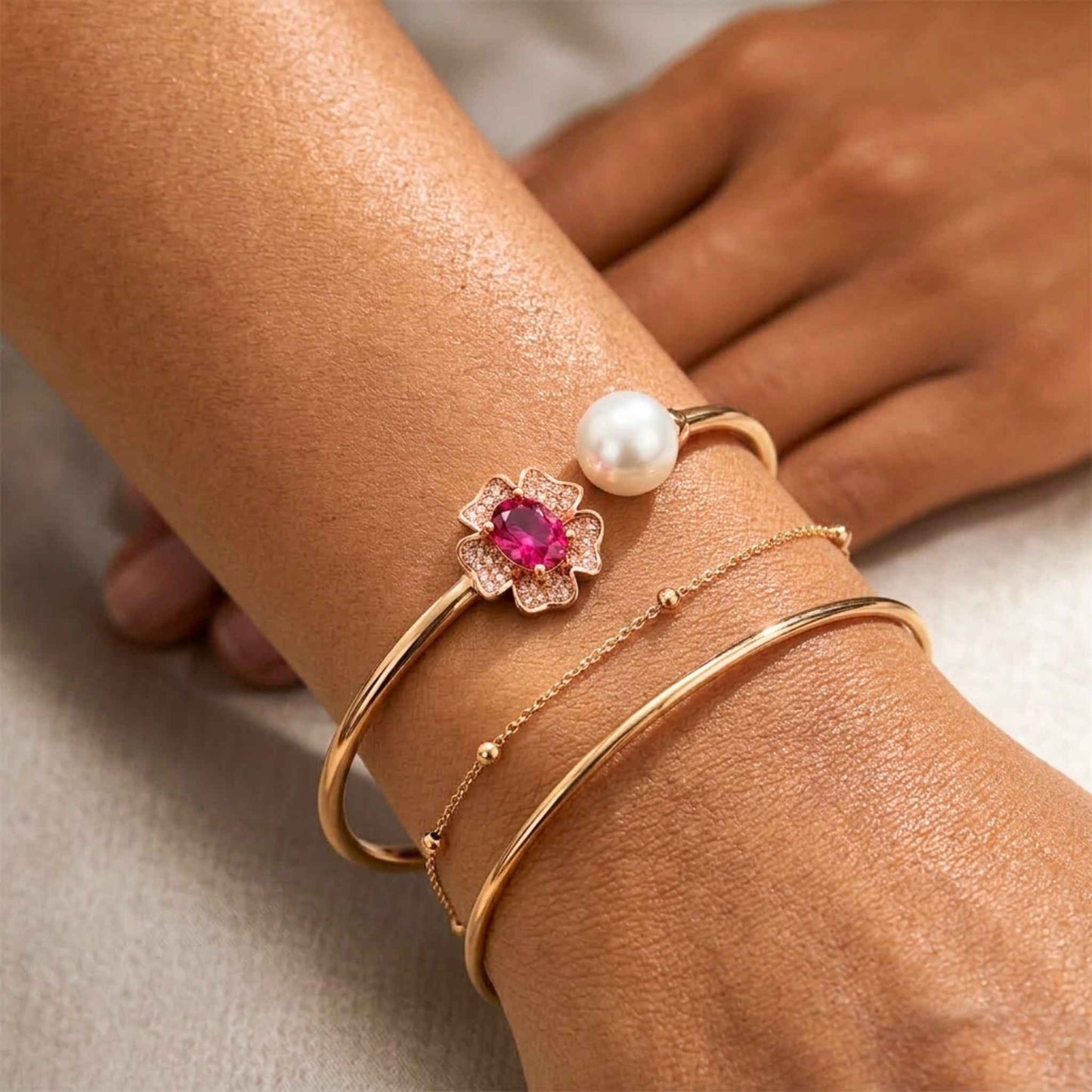 Close-up of a wrist wearing gold bracelets with a central ruby red gemstone and pearl on a natural background