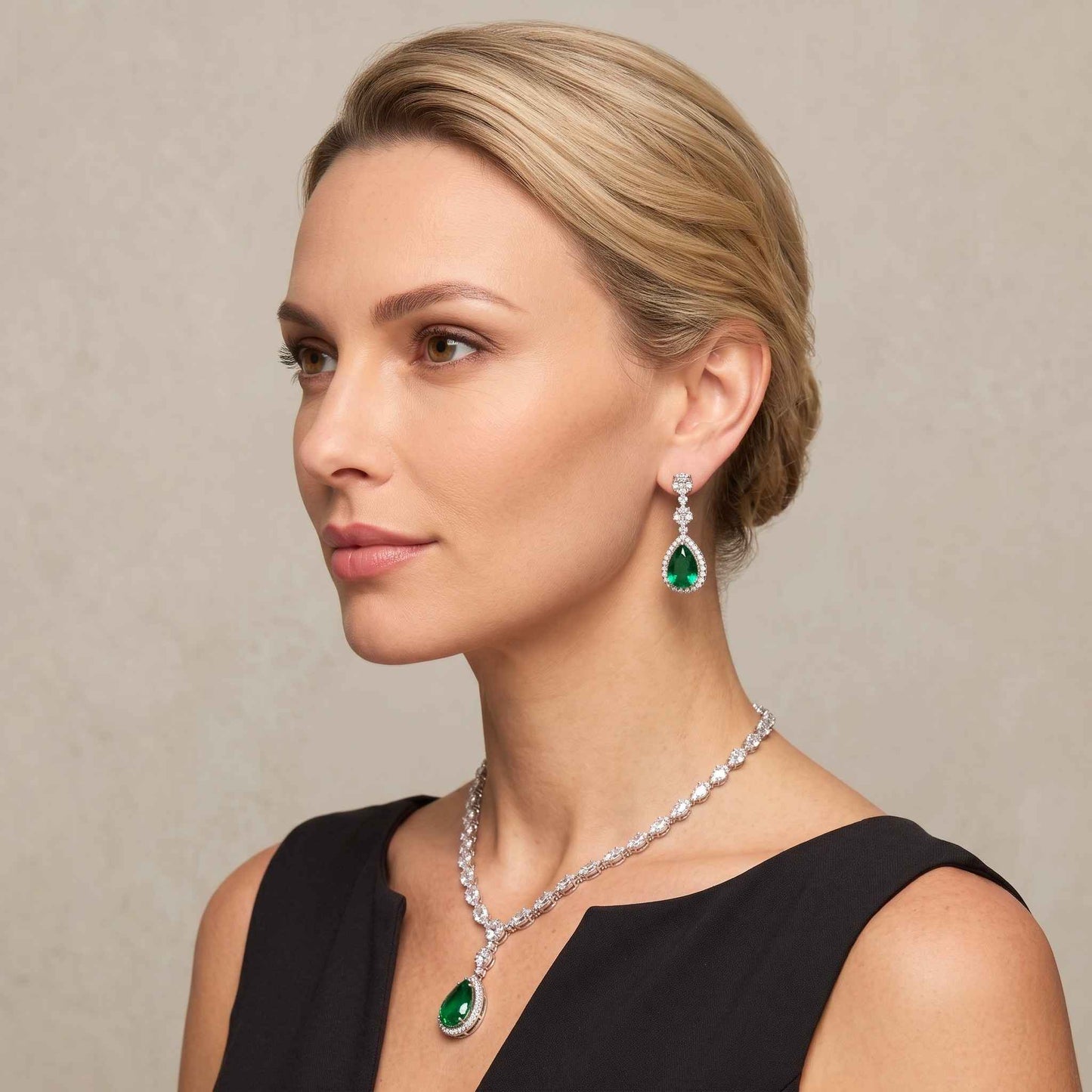 A woman with blonde hair styled in an updo wears a matching emerald green and pear shape gemstone necklace and drop earrings against a neutral beige background. Digitally rendered model for scale.