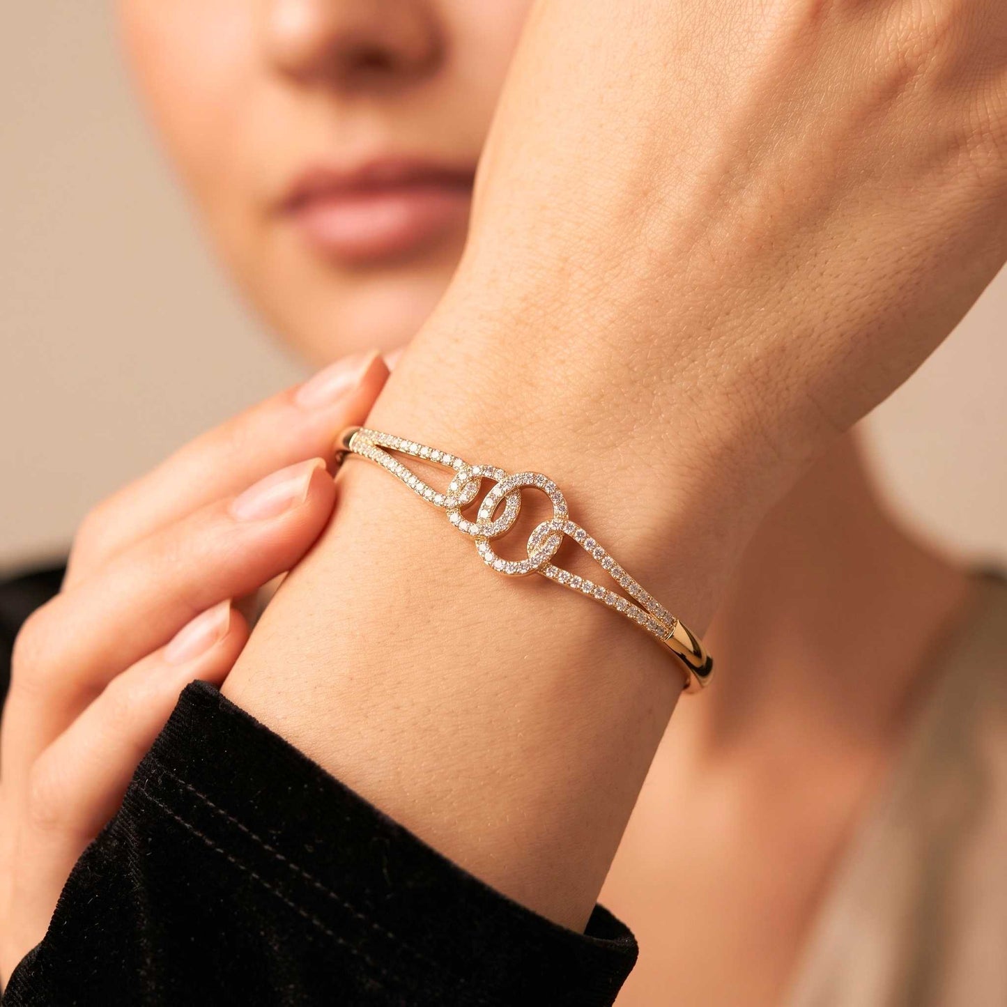 A close-up of a person's wrist wearing a delicate gold bangle bracelet adorned with pavé-set clear stones and a central interlocking double-O design. Digitally rendered model for scale