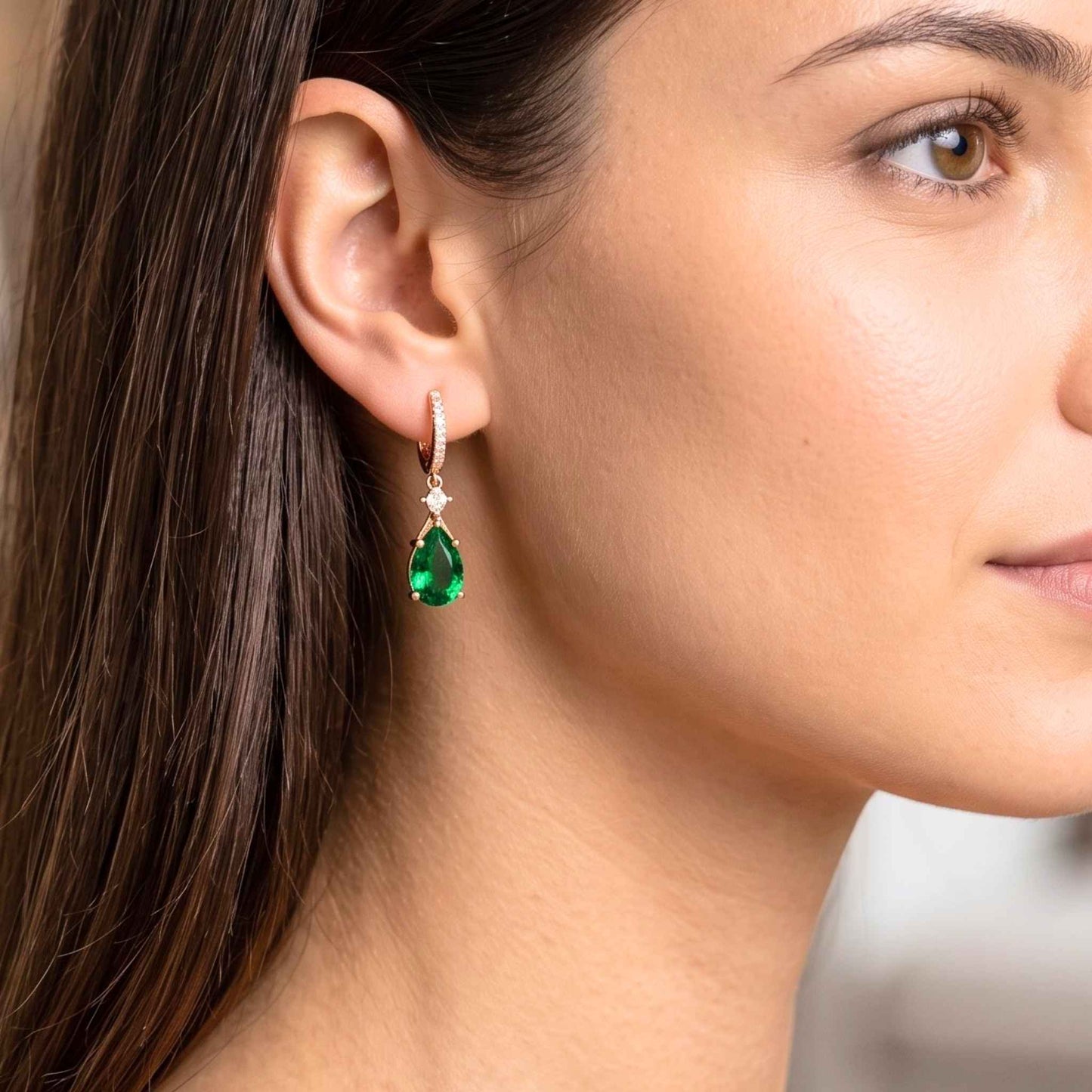 A close-up of a woman's ear wearing a single drop earring with a bright green teardrop gemstone and small clear stones above it in a gold-toned setting. Digitally rendered model for scale