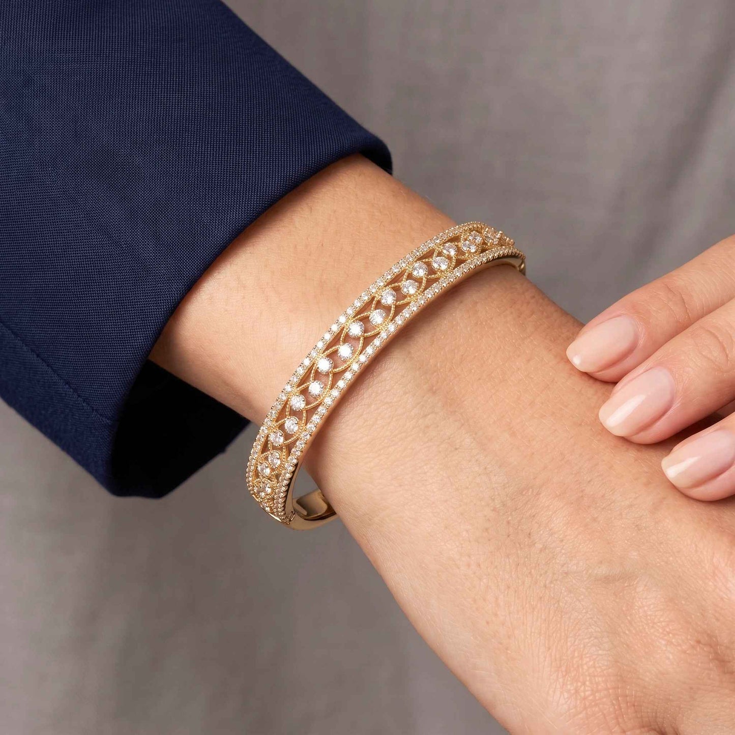 A close-up of a gold lattice bangle bracelet with marquise and round cubic zirconia, openwork detailing and a discreet concealed box clasp, worn on a person's wrist, with a navy blue sleeve visible and a neutral grey background. Digitally rendered model for scale