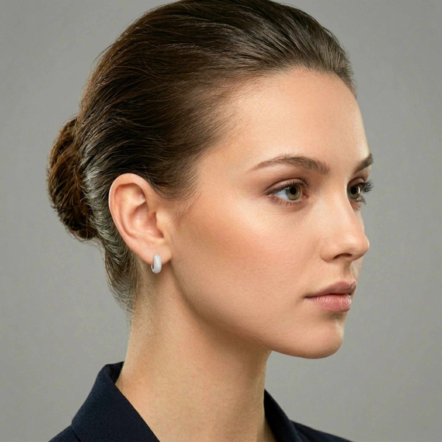 Side view of a woman's face who is wearing white gold plated huggie hoop earrings featuring rows of shimmering pavé-set clear cubic zirconia gemstones from Jewellery 4 Elegance collection set against a neutral background. Digitally rendered model for scale