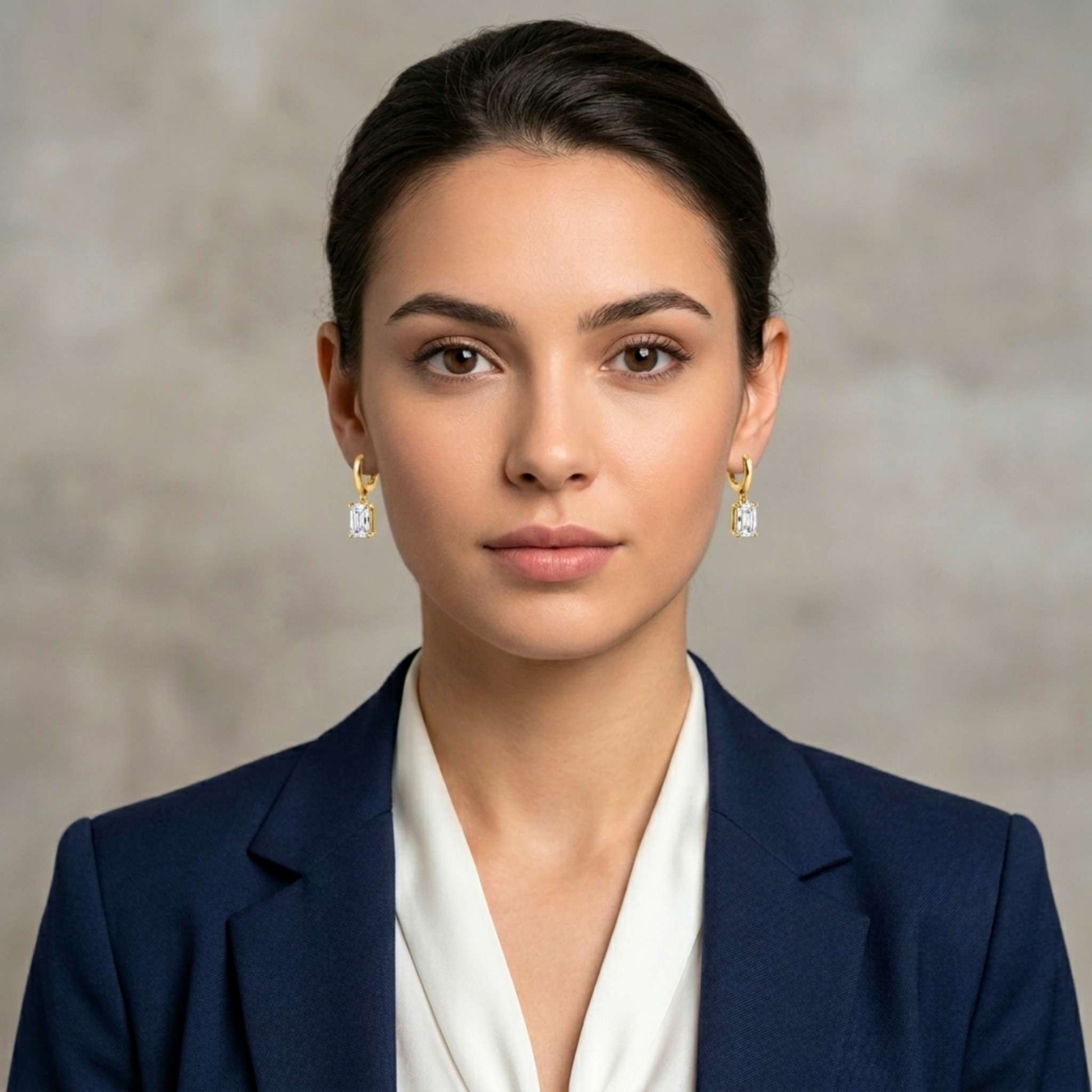 Woman wearing a pair of Jewellery 4 Elegance gold plated drop earrings featuring smooth huggie hoops suspending a brilliant emerald cut cubic zirconia gemstone and a navy blazer and white shirt against a neutral background.  Digitally rendered model for scale