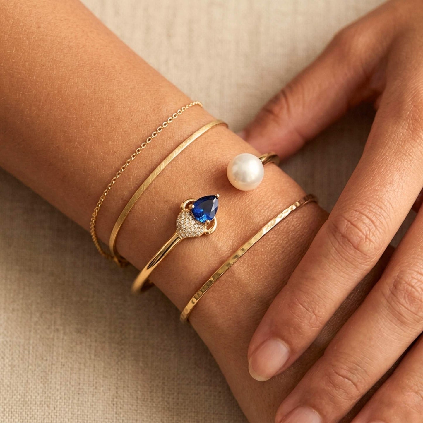 Close-up of a hand wearing multiple gold bracelets with a blue gemstone and pearl on a beige background.  Digitally rendered person wearing authentic bangle bracelet jewellery.