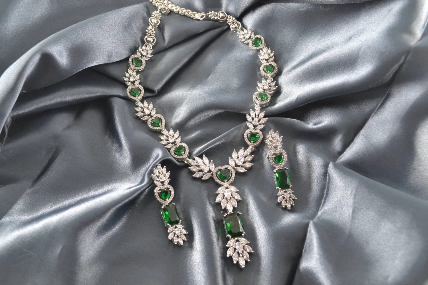 White gold necklace and earrings with green gemstones on grey satin fabric.