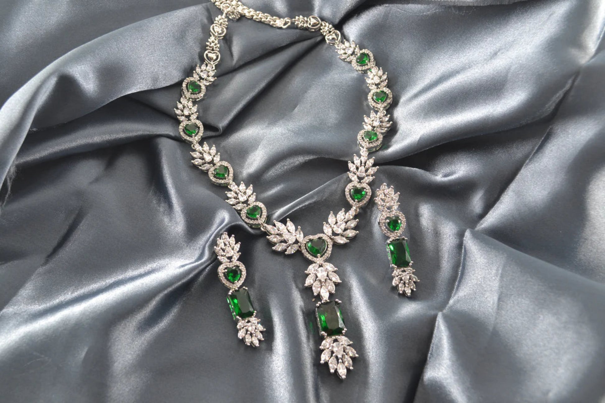 White gold necklace and earrings with green gemstones on grey satin fabric.