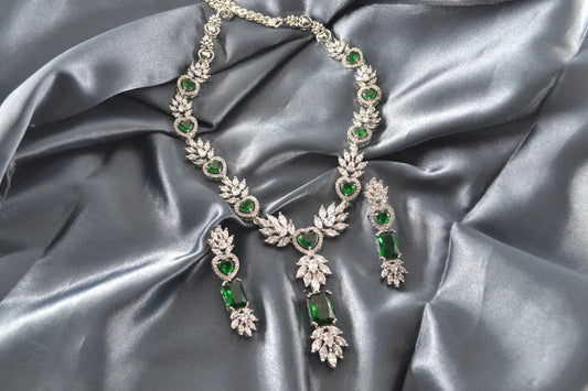 White gold necklace and earrings with green gemstones on grey satin fabric.