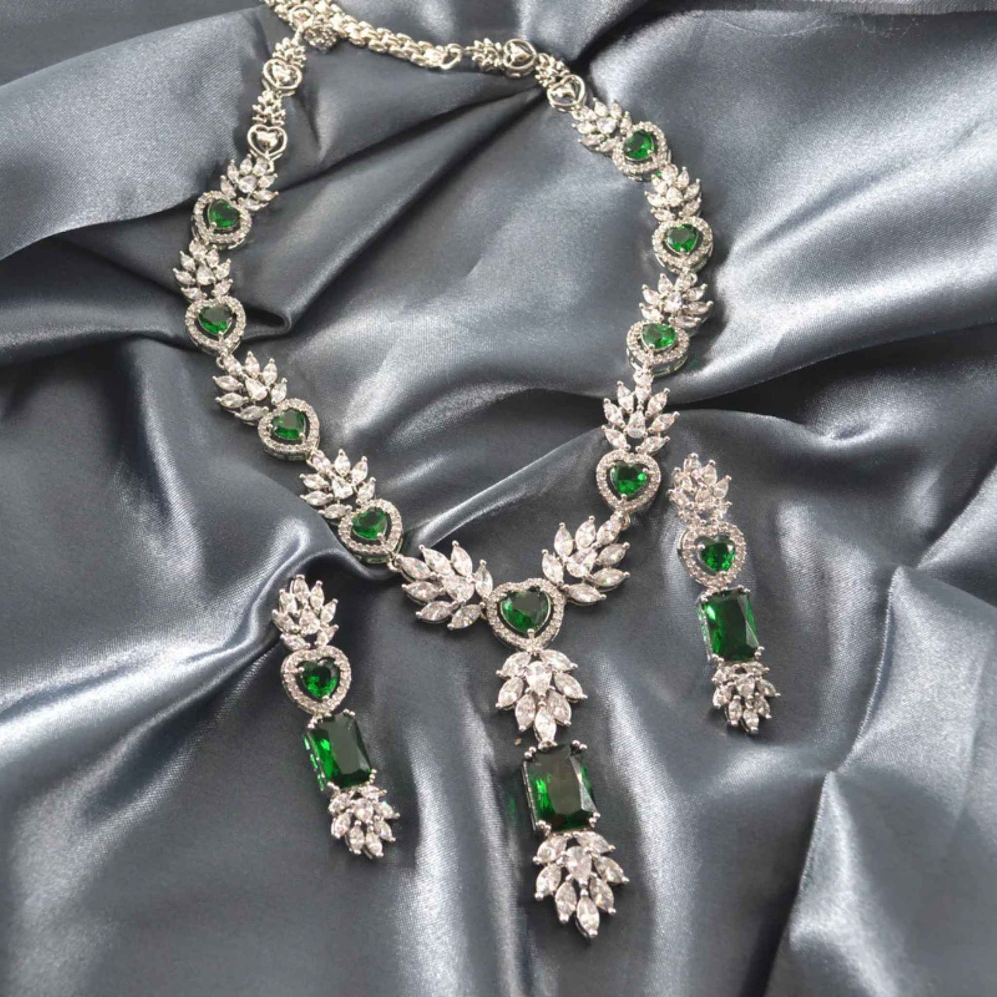 White gold necklace set with white and emerald green gemstones with matching earrings laying on a grey satin cloth.