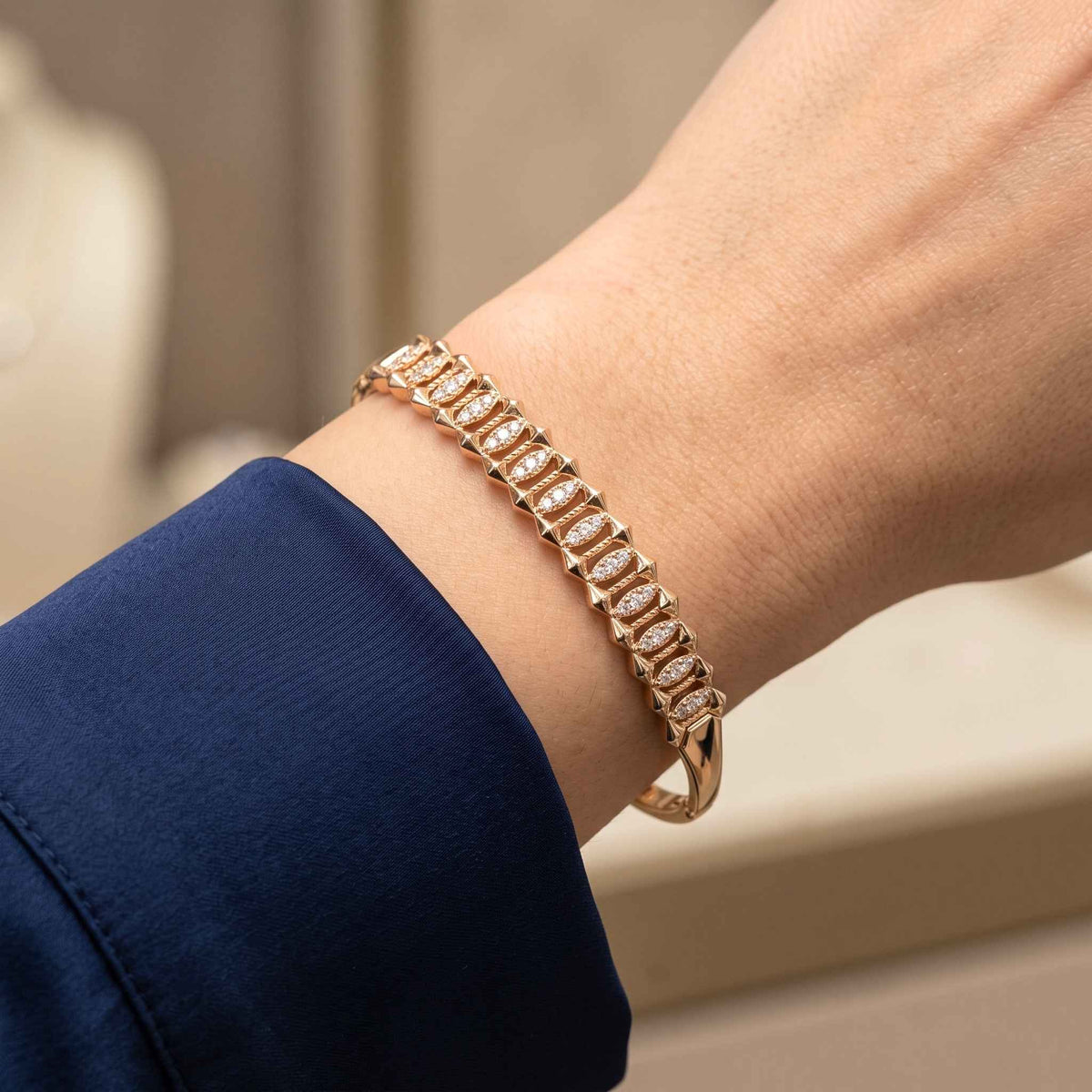 A close-up of a gold bangle bracelet from the Jewellery 4 Elegance collection featuring a geometric patter with stones, worn on a wrist with a navy blue sleeve visible.  Digitally rendered model for scale