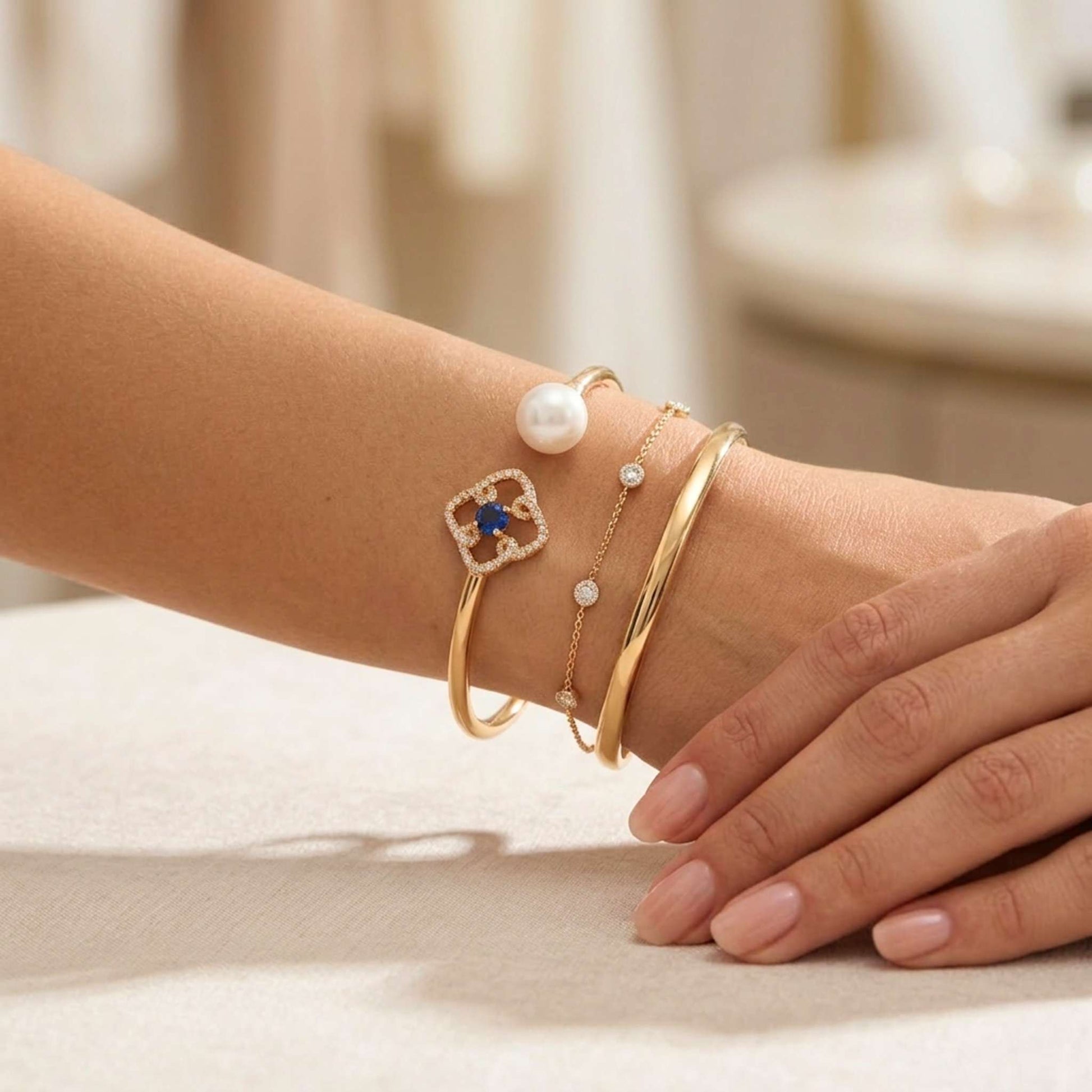 Close-up of a wrist wearing multiple gold bracelets with pearls and gemstones on a neutral background.