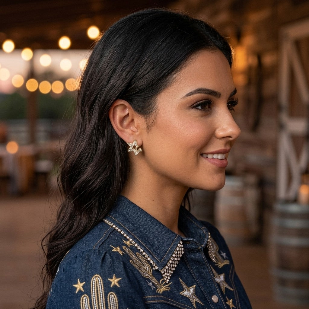 A woman in profile wearing a denim western-style jacket with gold embroidered stars and cactus details, along with matching star-shaped earrings, set against a warm, rustic barn interior with string lights.  Digitally rendered model for scale.