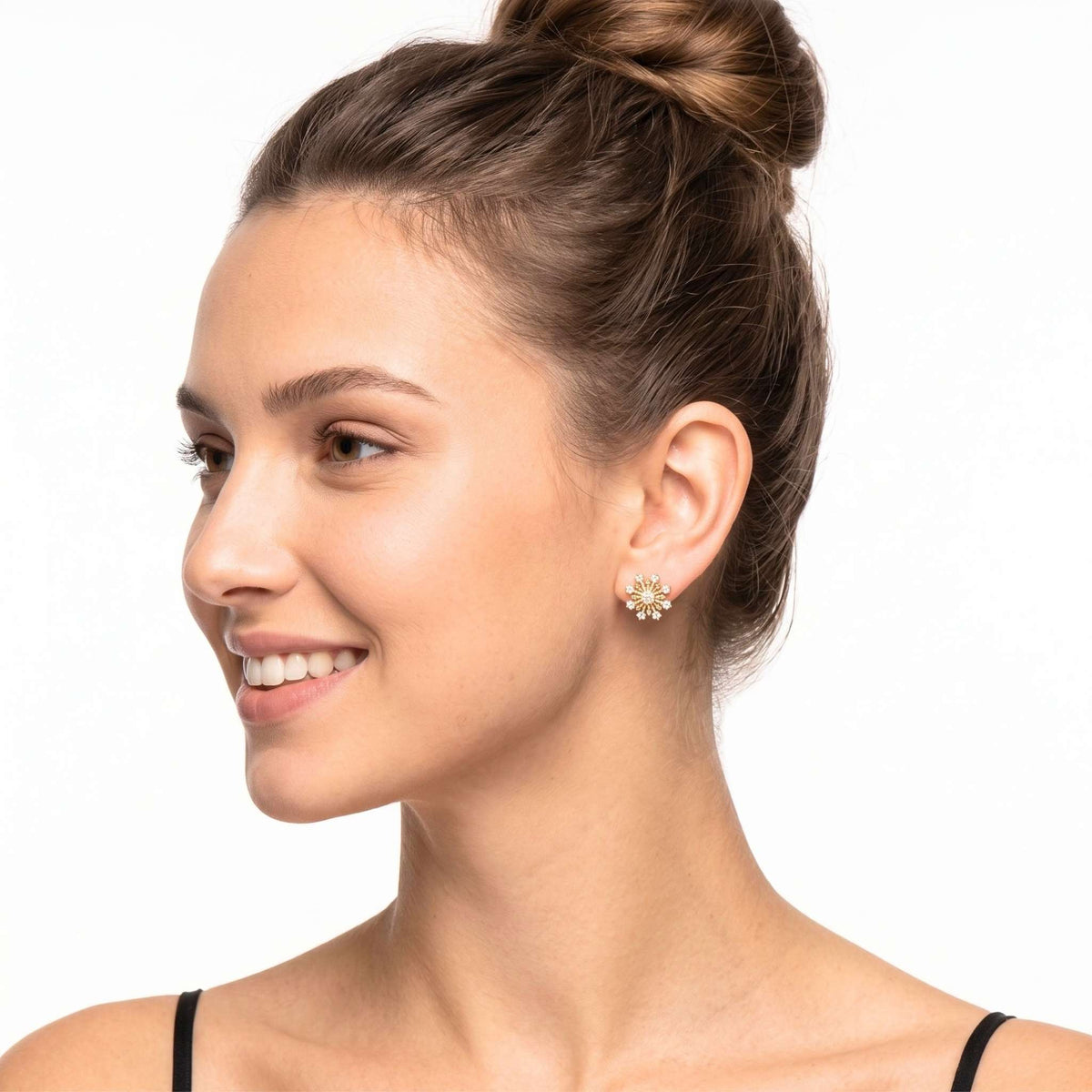 A woman with her hair in a bun is shown in profile wearing a small gold flower-shaped stud earring with clear stones from the Jewellery 4 Elegance collection, on a plain white background.   Digitally rendered model for scale