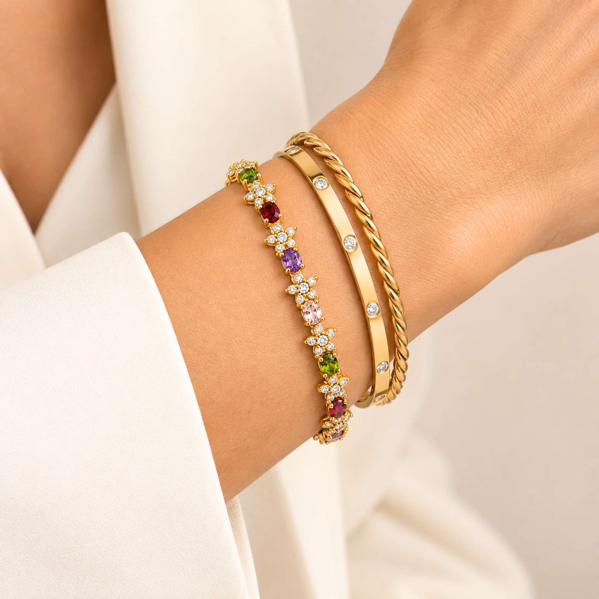 Three gold bracelets worn on a wrist: a thin bangle with small clear stones, a twisted bangle, and a floral bangle and a multi-coloured bracelet from the Jewellery 4 Elegance collection featuring gemstones including green, purple, red, and pink. The wrist is shown against a soft white garment background.  Digitally rendered model for scale