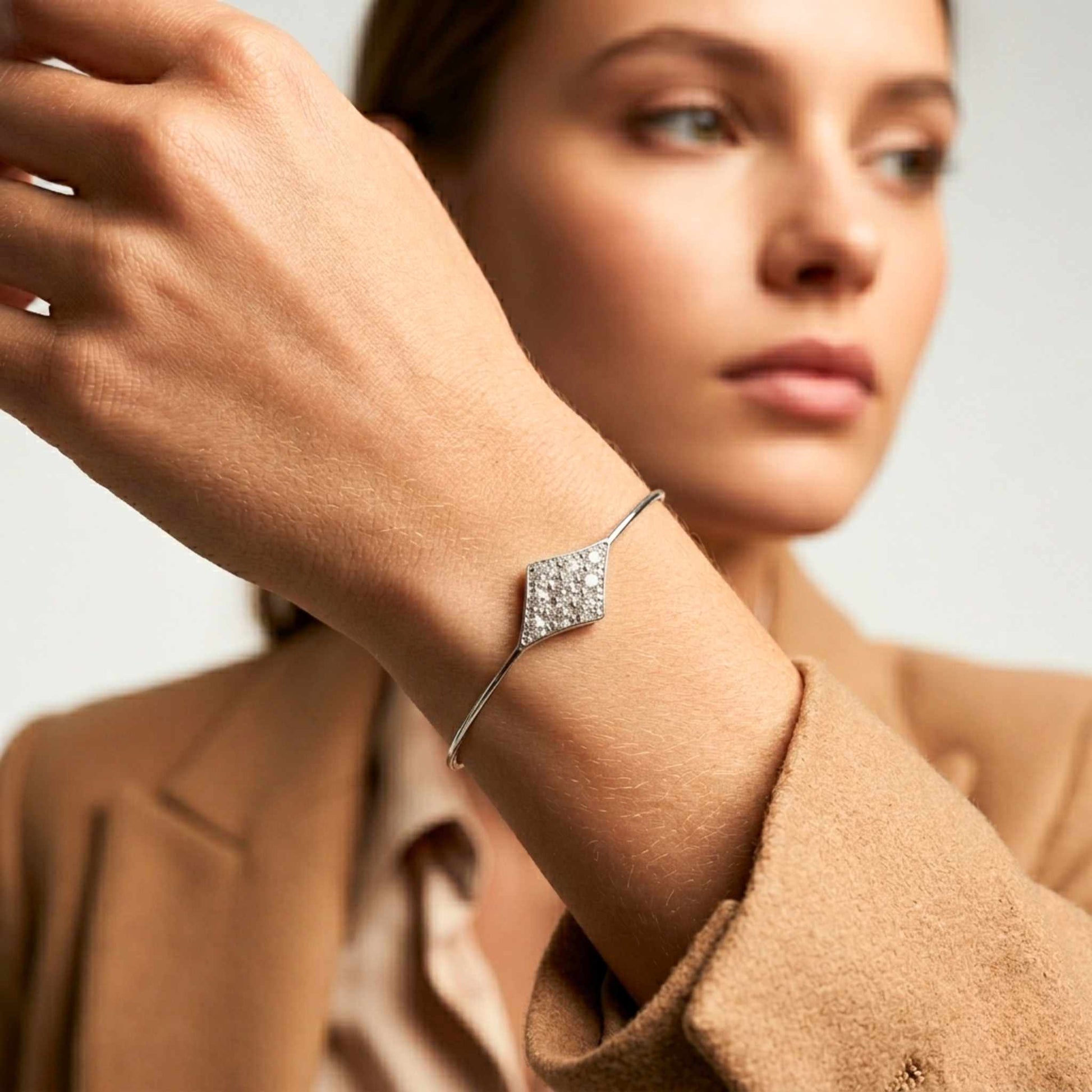 Woman in a brown jacket wearing a white gold bangle bracelet featuring a central diamond-shaped motif packed with sparkling pavé-set cubic zirconia stones from Jewellery 4 Elegance collection.  Digitally rendered model for scale.