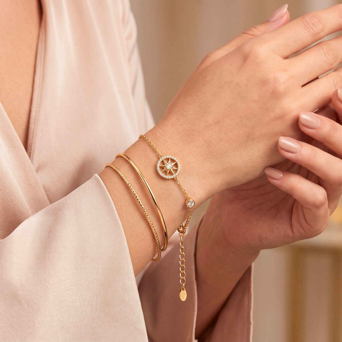 A close-up of a person's wrist and hand wearing delicate gold bracelets, including a starburst charm bracelet from the Jewellery 4 Elegance collection, a thin chain bracelet, and a bangle, styled with a soft beige satin robe.  Digitally rendered model for scale