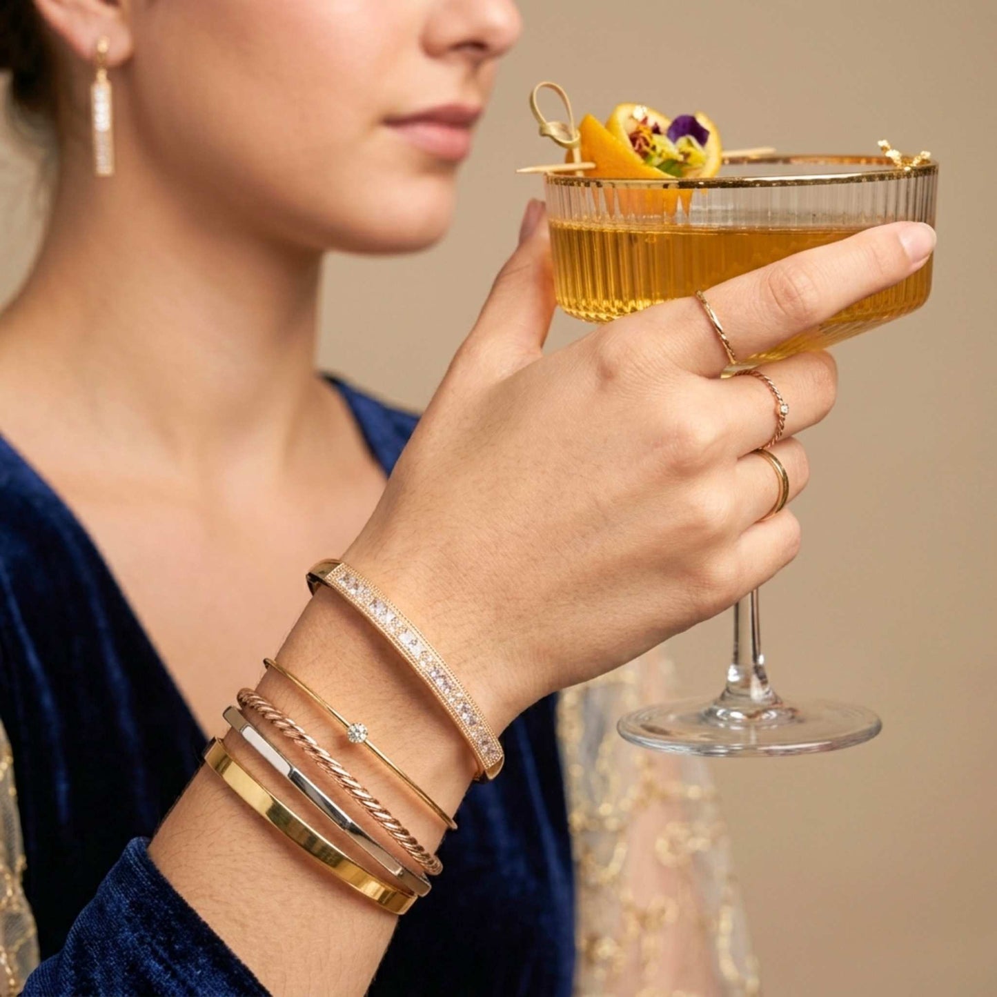 Woman holding a cocktail with gold jewellery on a neutral background showing a bangle bracelet from the Jewellery 4 Elegance.  Digitally rendered model for scale.