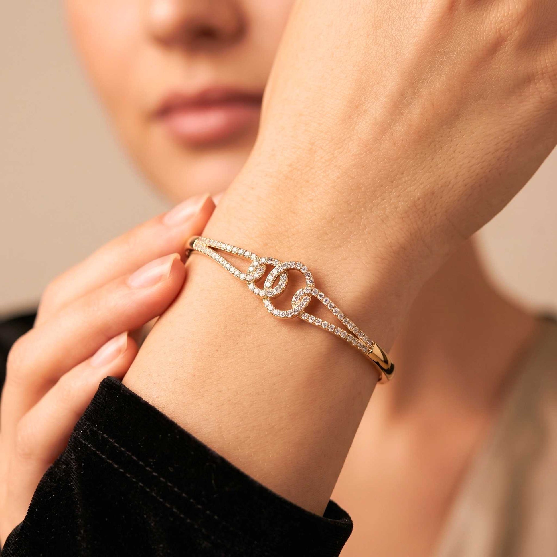 A close-up of a person's wrist wearing a delicate gold bangle bracelet adorned with pavé-set clear stones and a central interlocking double-O design. Digitally rendered model for scale