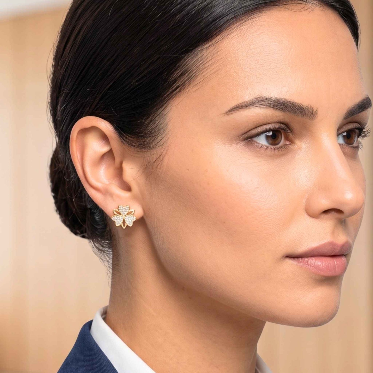 A professional portrait of a woman with dark hair wearing a navy blue blazer modelling a pair of gold stud earrings in a sweet clover design featuring cubic zirconia from the Jewellery 4 Elegance set against a softly blurred office background.  Digitally rendered model for scale