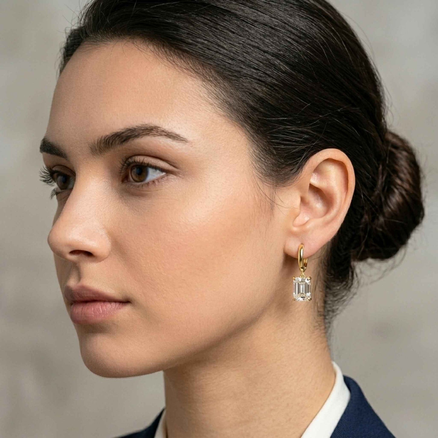 Close-up of a woman wearing a pair of Jewellery 4 Elegance gold drop earrings featuring smooth huggie hoops suspending a brilliant emerald cut cubic zirconia gemstone and a navy blazer and white shirt against a neutral background.  Digitally rendered model for scale