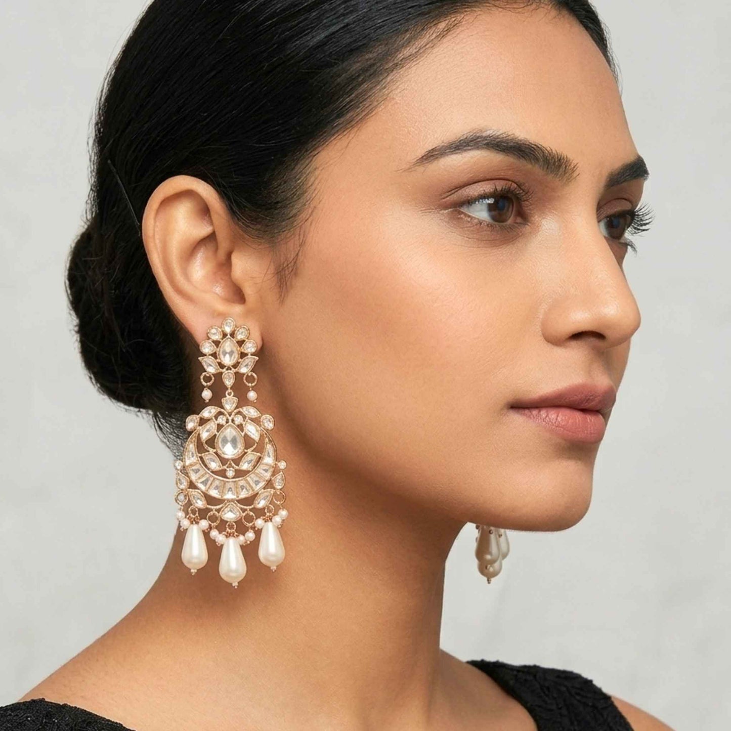 Woman wearing a black dress with gold Indian Chandbali chandelier drop earring featuring intricate cubic zirconia encrusting and dangling teardrop pearl beads from Jewellery 4 Elegance set against a neutral background.  Digitally rendered model for scale