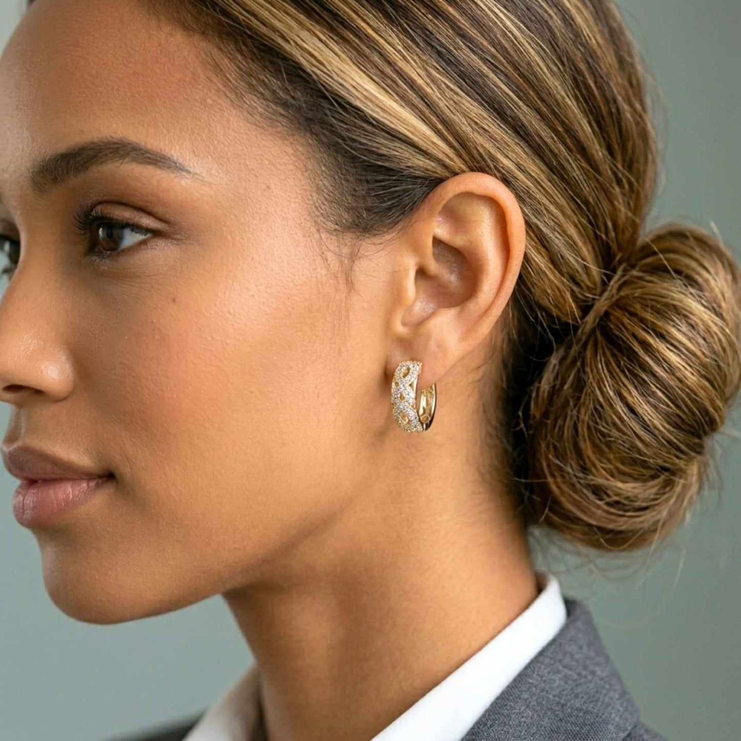Woman wearing pair of Jewellery 4 Elegance gold plated small hoop earrings featuring an openwork infinity latticework design set with pavé cz gemstones. and white shirt with a neutral background.  Digitally rendered model for scale
