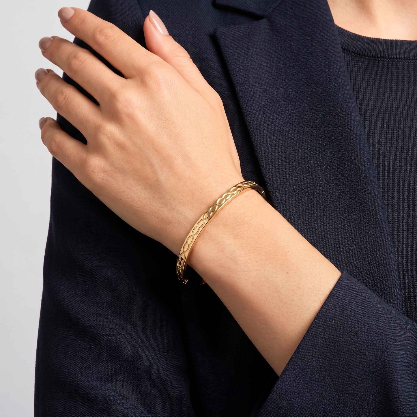 A close-up of a person's hand wearing a gold infinity link engraved bangle bracelet with a discreet concealed box clasp from the Jewellery 4 Elegance collection.  Digitally rendered model for scale
