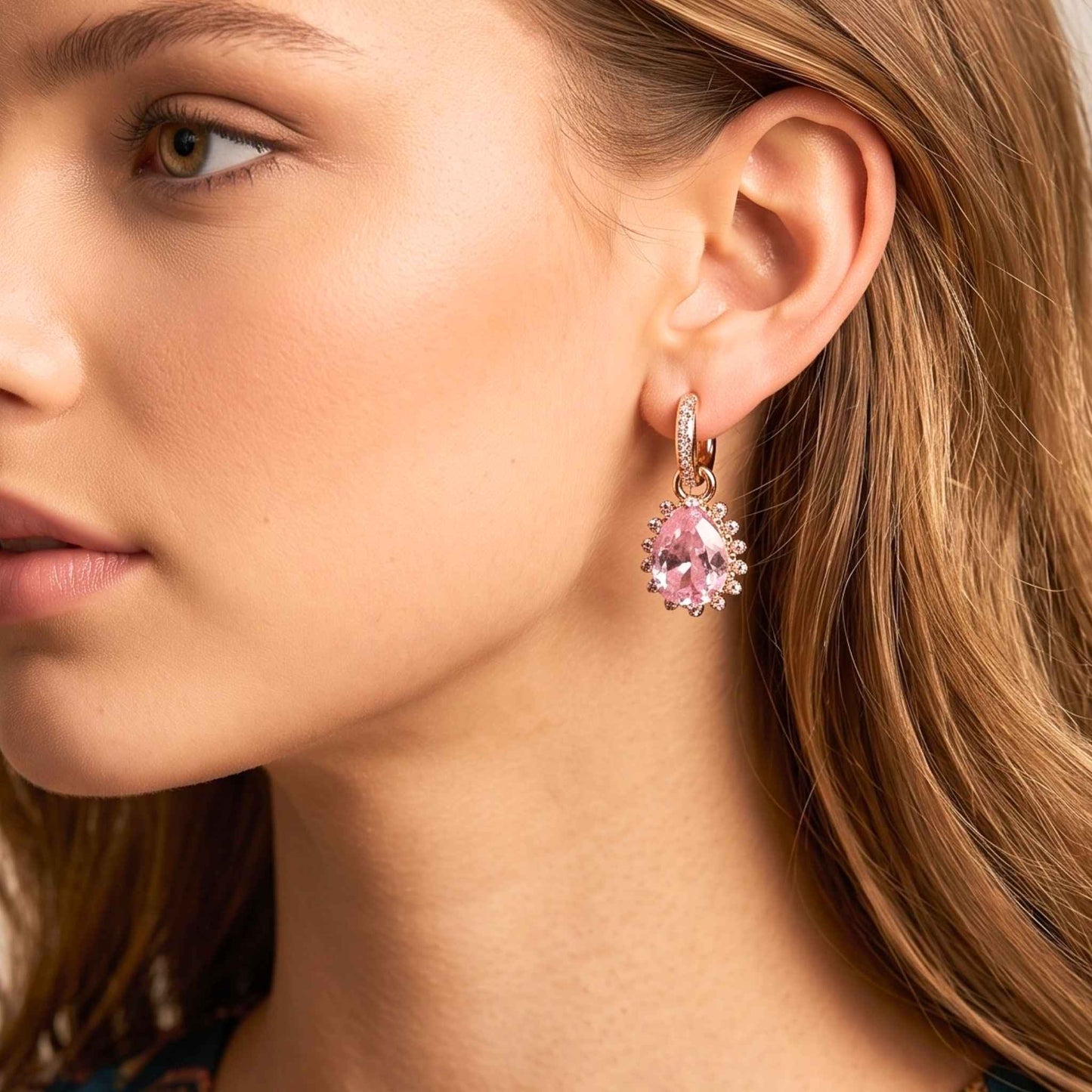 Close up of a woman with wavy light brown hair wearing gold plated hinged hoop drop earrings featuring faceted pink pear-cut stones surrounded by a delicate cubic zirconia halo with a polished gold finish set against a plain background.  Digitally rendered model for scale.