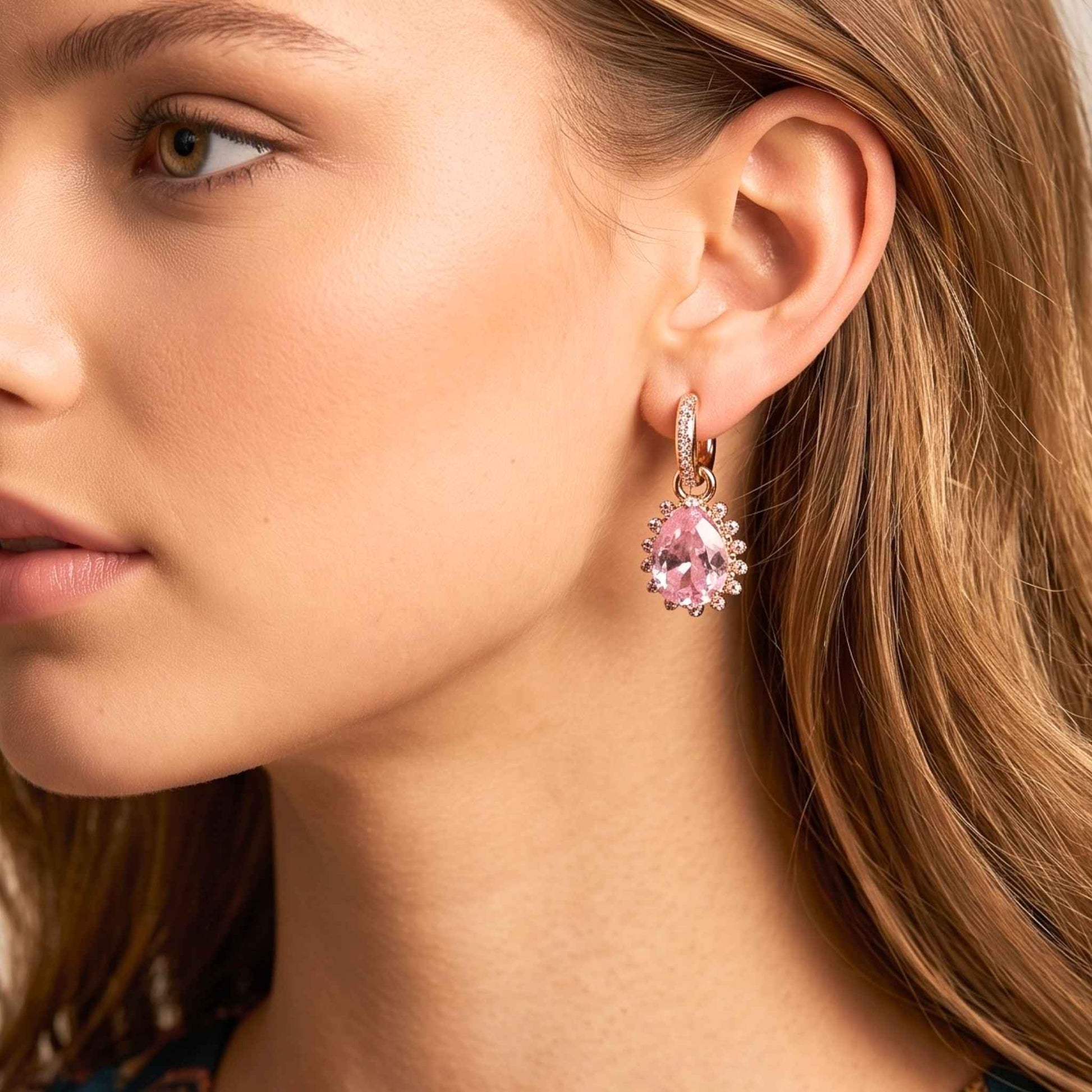 Close up of a woman with wavy light brown hair wearing gold plated hinged hoop drop earrings featuring faceted pink pear-cut stones surrounded by a delicate cubic zirconia halo with a polished gold finish set against a plain background.  Digitally rendered model for scale.