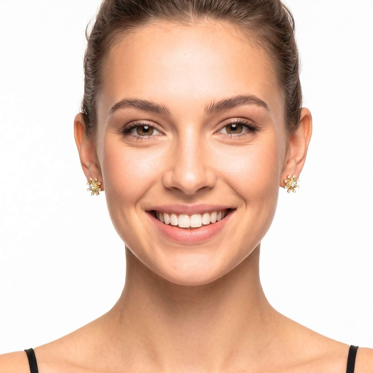 A smiling woman with her hair pulled back wears small gold snowflake shaped stud earrings with clear stones from the Jewellery 4 Elegance collection, against a plain white background.  Digitally rendered model for scale