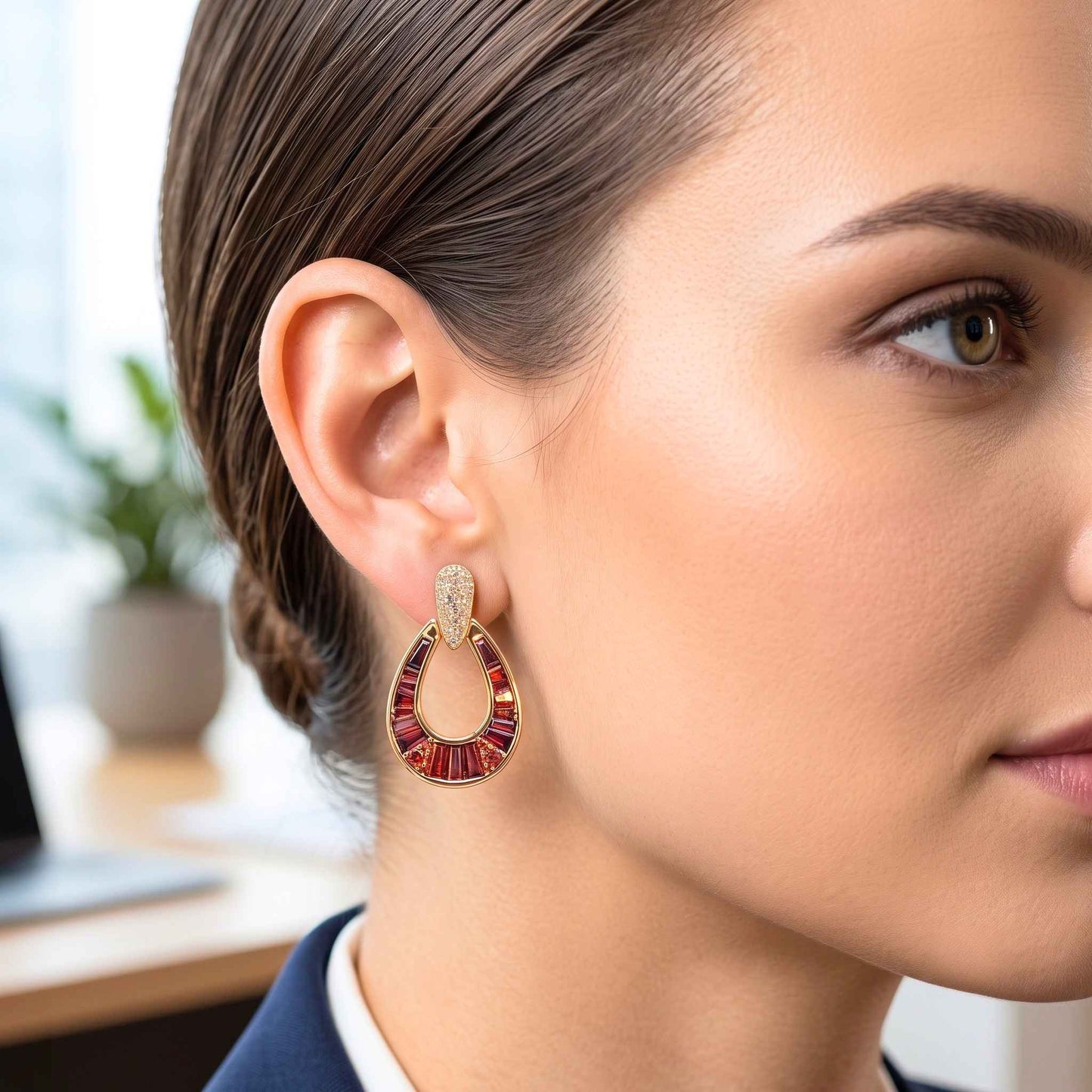 Close up of a woman with neatly styled brown hair wearing a navy blazer and white collared shirt, showcasing a pair of red and gold horseshoe drop earring featuring garnet red baguette-cut stones with pavé cubic zirconia for an elegant contemporary look from the jewellery 4 elegance collection. Digitally rendered model for scale.