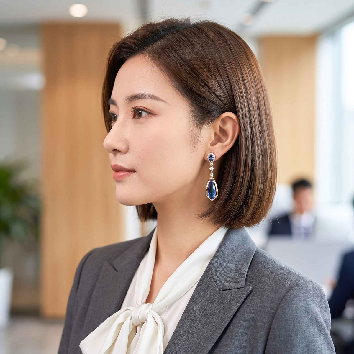 A woman in a grey blazer and cream blouse wearing a dangling teardrop earring with a blue stone and silver-toned setting, photographed in a softly blurred office interior.  Digitally rendered model for scale