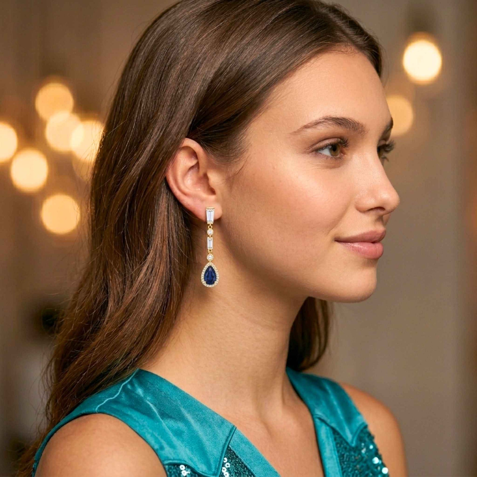 Gold plated drop earrings featuring pear-shaped sapphire blue cubic zirconia stones with pavé halo detailing and baguette-cut cubic zirconia accents. Digitally rendered model for scale