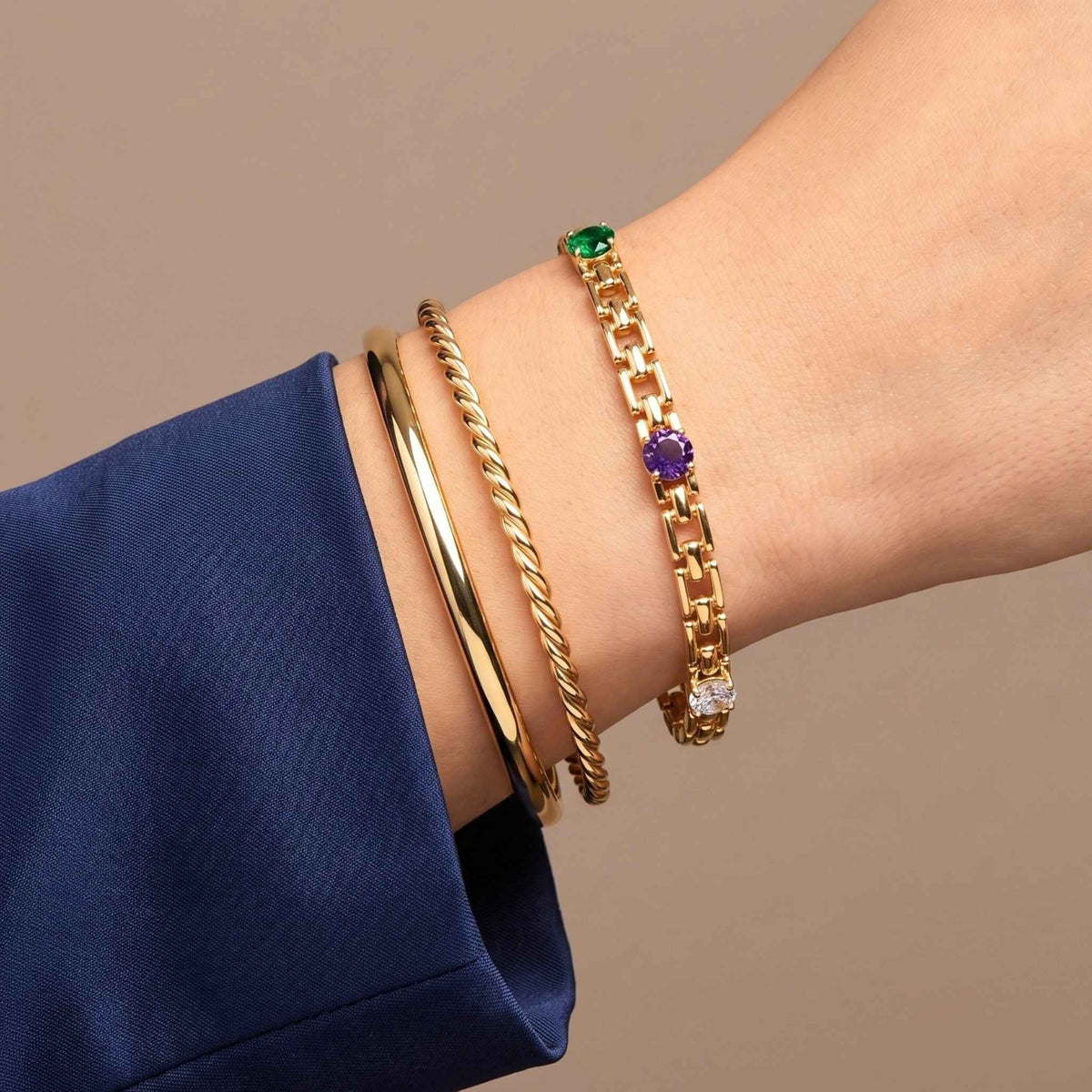 A close-up of a person's wrist wearing three gold bracelets: a plain gold bangle, a twisted gold bangle, and a gold link bracelet with a green, purple and clear gemstone accents from the Jewellery $ Elegance collection.  Digitally rendered model for scale