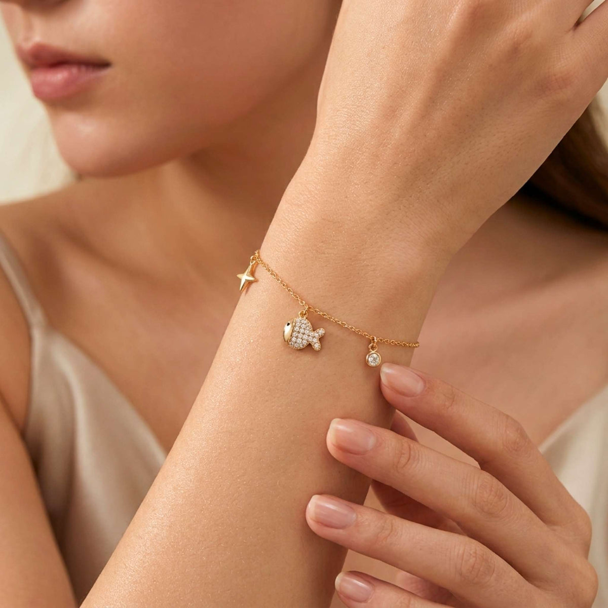 A delicate gold chain bracelet with a small fish charm, a star charm, and a tiny pearl-like bead, shown worn on a woman's wrist.  Digitally rendered model for scale