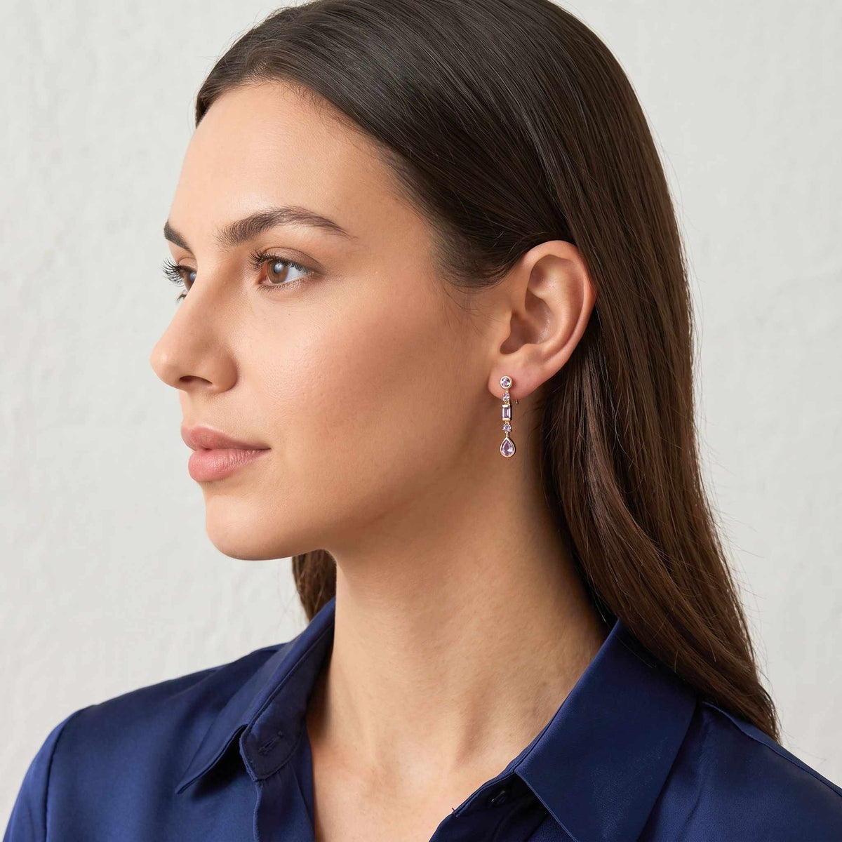 A close-up portrait of a woman wearing  purple gemstone drop earrings with teardrop-shaped drops, paired with a navy blue collared shirt against a light grey background.  Digitally rendered model for scale