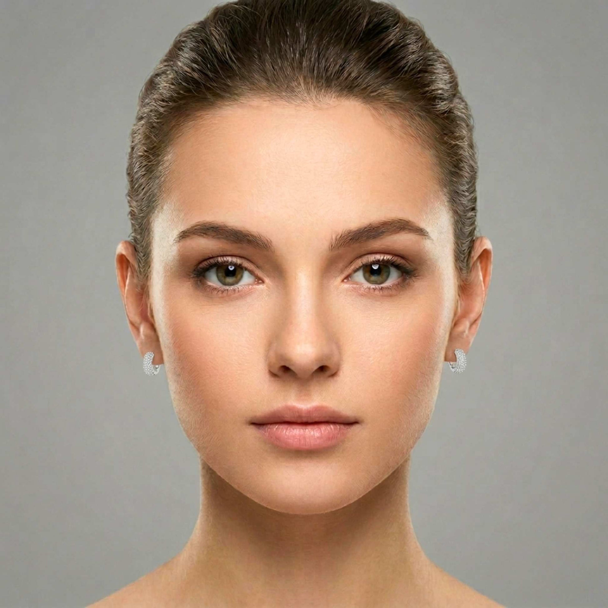 Close-up of a woman's face who is wearing white gold plated huggie hoop earrings featuring rows of shimmering pavé-set clear cubic zirconia gemstones from Jewellery 4 Elegance collection set against a neutral background.  Digitally rendered model for scale