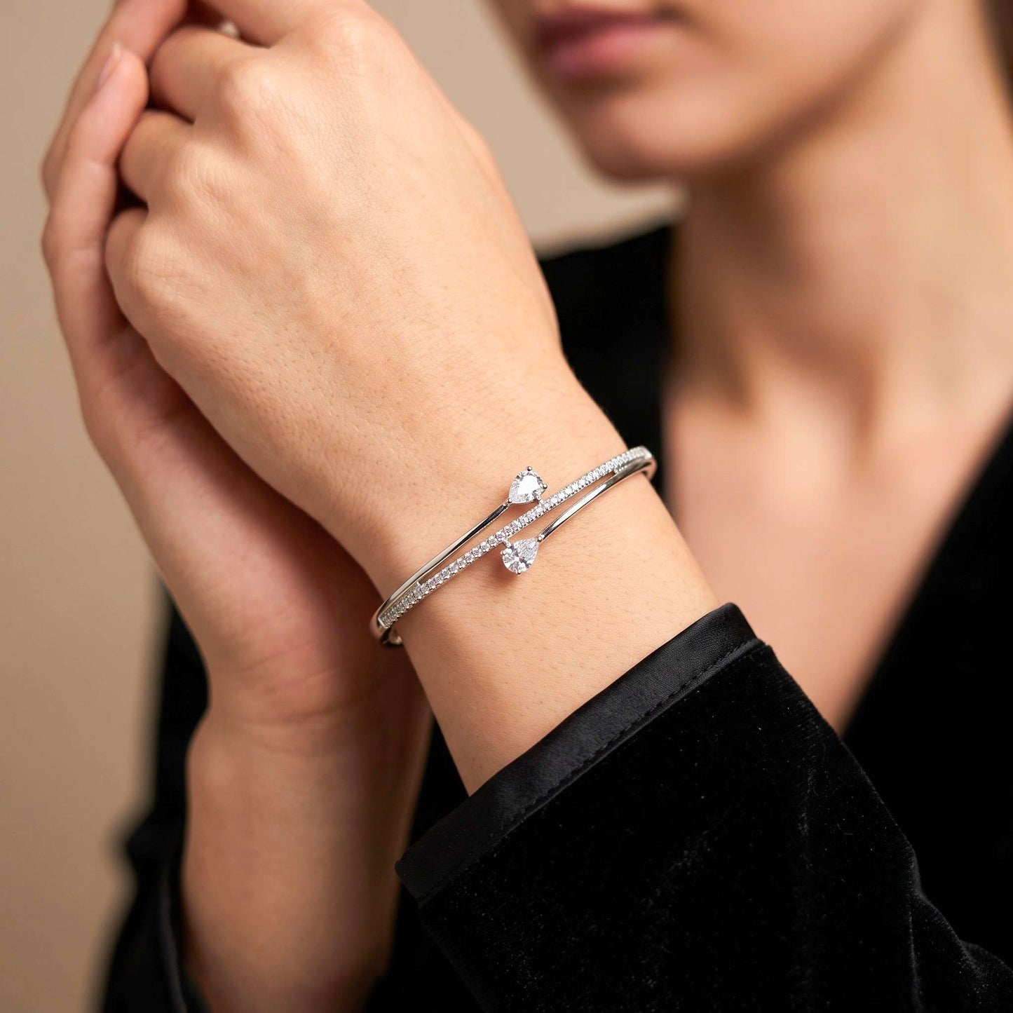 A close up of a delicate white gold crossover bangle bracelet with two pear-cut cubic zirconia, a row of round cubic zirconia and a concealed clasp, paired with a black velvet top.  Digitally rendered model for scale
